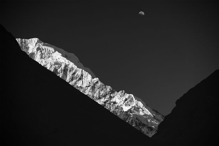 Dramatic minimalist photography of snow-capped mountains in black and white, with a crescent moon in the dark sky.