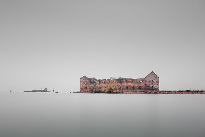 Extraordinary minimalist photography of ancient ruins on a small island surrounded by calm water under a gray sky.