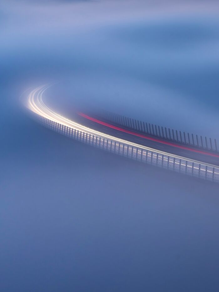 A minimalist photography award winner, featuring streaks of car lights curving through dense fog on a bridge, creating an abstract glow.