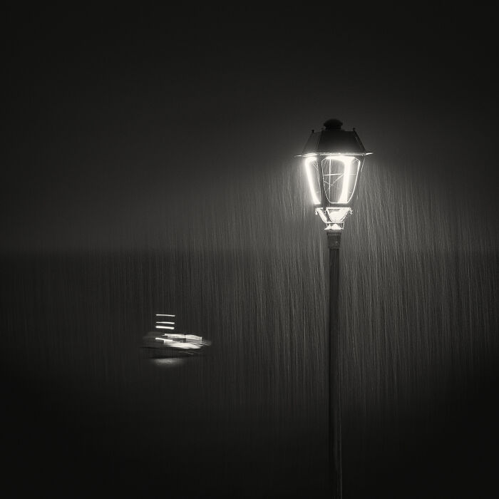 A glowing street lamp illuminates rain falling in a dark, minimalist scene, captured for Minimalist Photography Awards.
