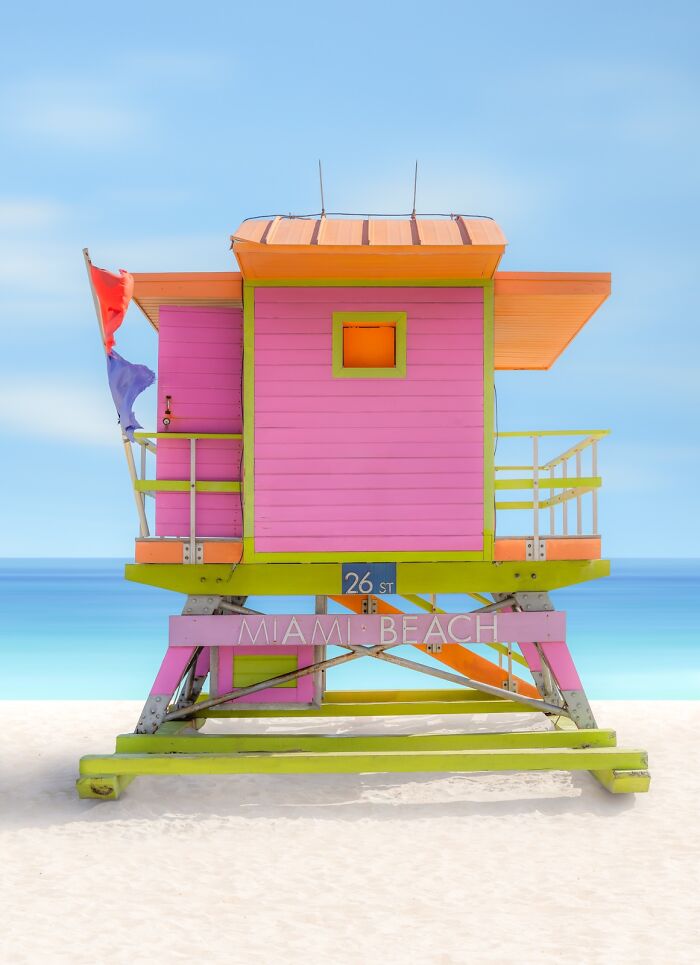 A vibrant pink and orange lifeguard stand on a white sand beach with a clear blue sky, demonstrating minimalist photography.