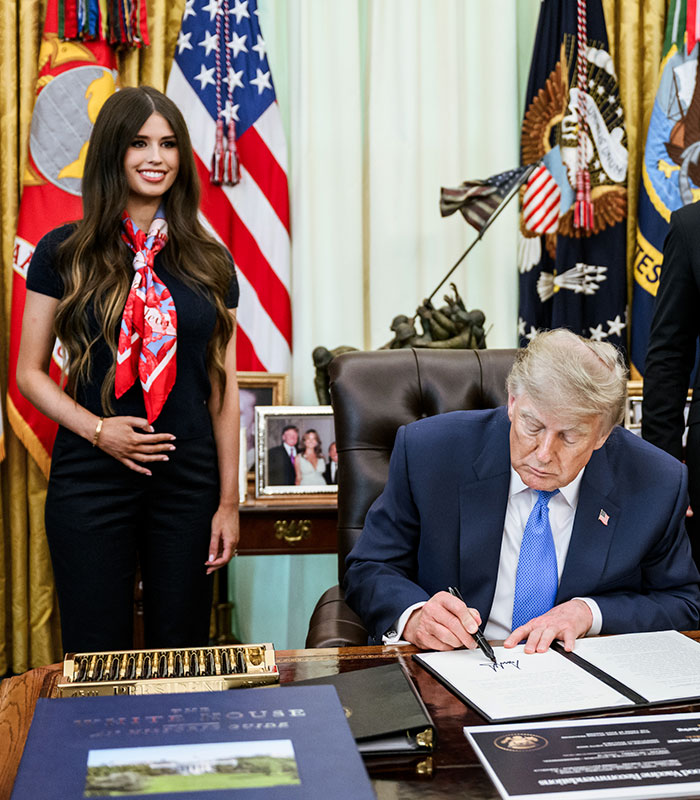Donald Trump signing documents while the mystery woman stands behind him with a strange gesture. Donald Trump signing documents while the mystery woman stands behind him with a strange gesture.