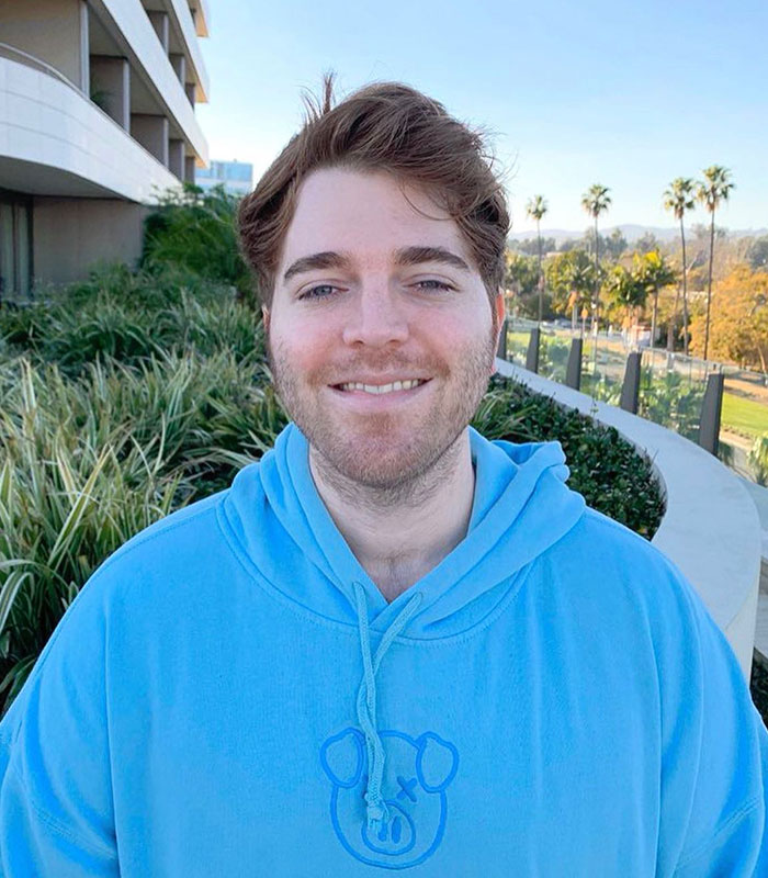 Influencers exposed for lying: A smiling man in a blue hoodie with a pig logo, outdoors with palm trees and a building.