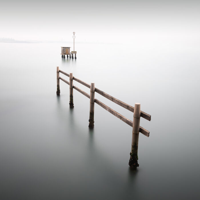 Minimalist photography of a wooden pier structure emerging from calm, misty water under a soft, white sky.
