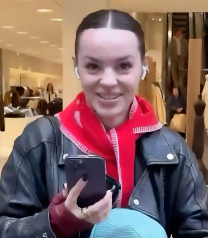 A daughter is surprised by her family and gives a slight smile, holding a phone, after their across-the-world surprise.