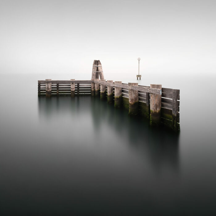 Minimalist photography of a wooden pier structure emerging from calm, misty water under a soft, white sky.