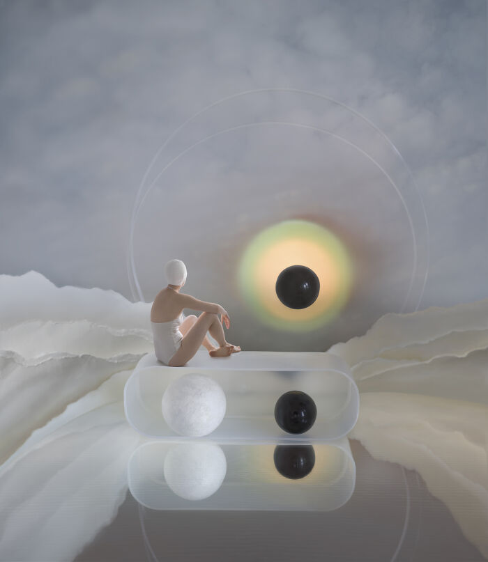 A minimalist photography award winner showing a person in white attire sitting on a transparent block, observing a floating black sphere.
