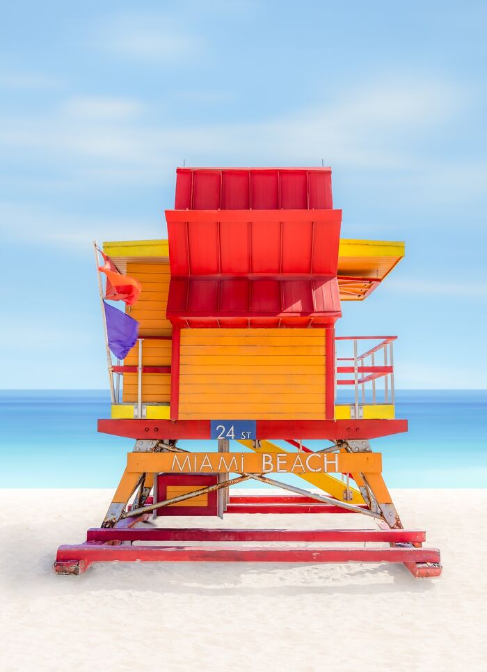 A vibrant pink and orange lifeguard stand on a white sand beach with a clear blue sky, demonstrating minimalist photography.