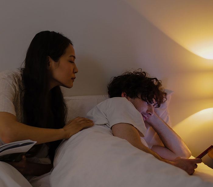 A woman looking concerned at her partner using his phone in bed, highlighting potential cheating.