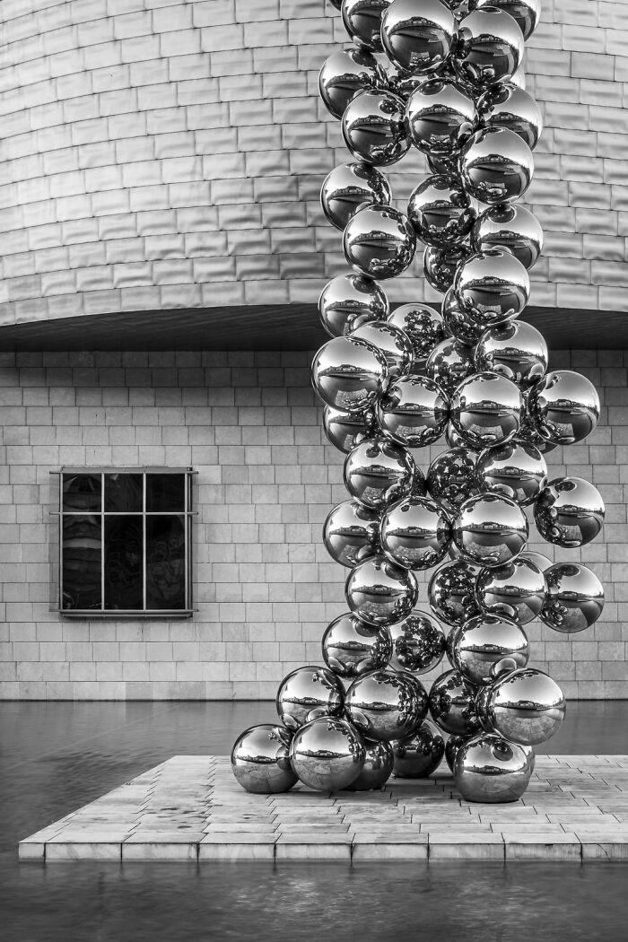 A striking black and white minimalist photography award-winning image of a large metallic spider sculpture in front of a modern, textured building.