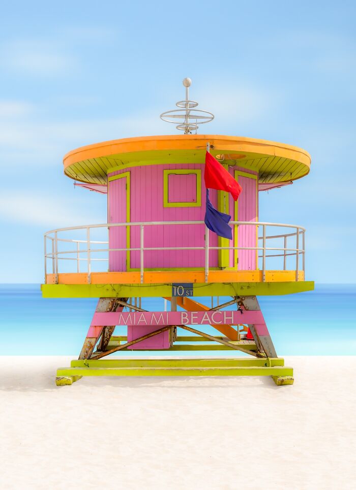 A vibrant pink and orange lifeguard stand on a white sand beach with a clear blue sky, demonstrating minimalist photography.