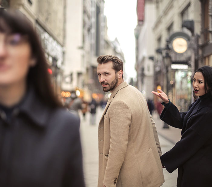 A man cheating and looking at another woman while his girlfriend holds his arm in a street.