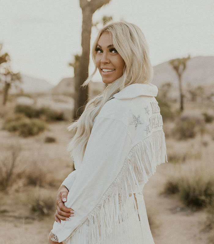Smiling blonde influencer in a fringed white jacket in a desert landscape, one of 11 influencers exposed for lying