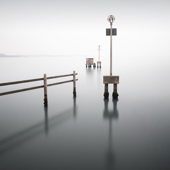 Minimalist photography of a wooden pier structure emerging from calm, misty water under a soft, white sky.