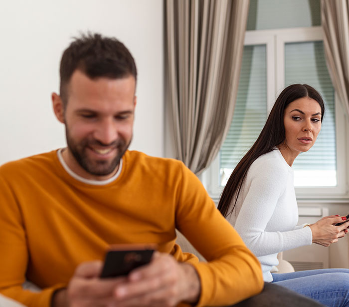 A woman looking suspiciously at her boyfriend, who is smiling while texting on his phone, hinting at cheating.