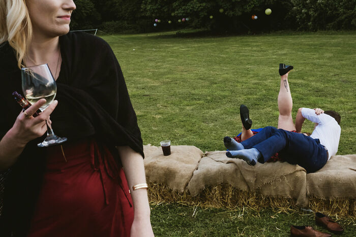 A woman holding a wine glass in the foreground, with two people lying on hay bales in the background, a humorous street photography scene.