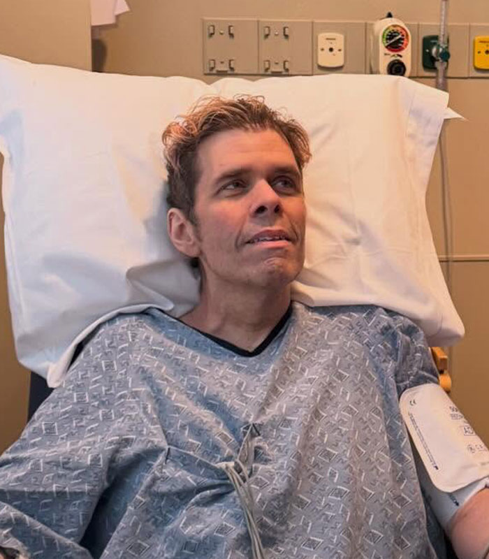Perez Hilton in a hospital gown, reclining in a bed, giving a firsthand account of his disturbing livestream.