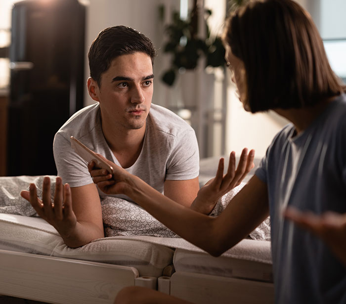 A couple in bed arguing about cheating, with the man holding his phone and the woman gesturing.