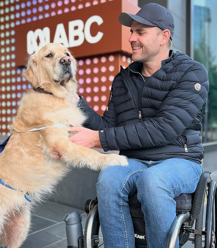 A smiling wheelchair advocate in a cap and black jacket, interacting with a service dog outside an ABC building. Netizens claim. A smiling wheelchair advocate in a cap and black jacket, interacting with a service dog outside an ABC building. Netizens claim.
