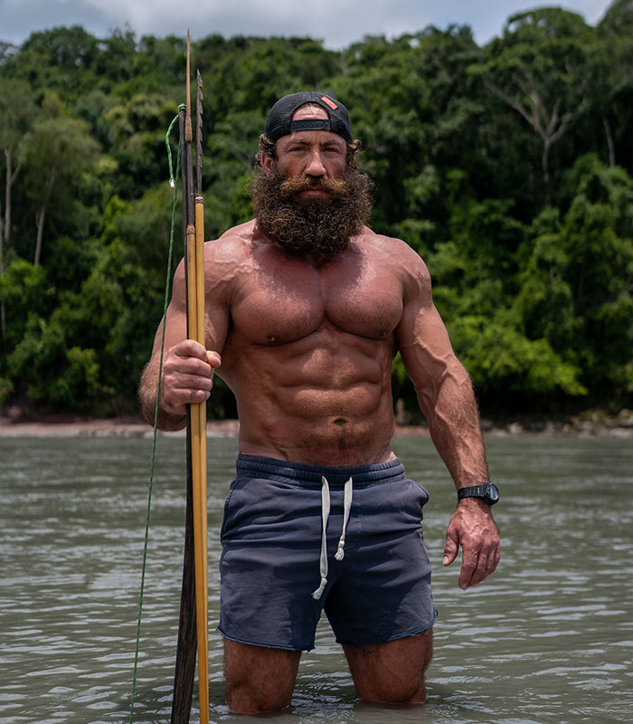 Influencers exposed for lying: A muscular man with a beard and spear standing in water with jungle in the background.
