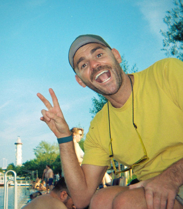 A man with a beard and cap, wearing a yellow shirt, smiling and giving a peace sign. Wheelchair advocate. A man with a beard and cap, wearing a yellow shirt, smiling and giving a peace sign. Wheelchair advocate.