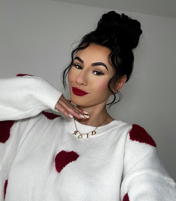 Influencers exposed for lying: A woman with dark hair in a bun, red lipstick, and a Dior necklace, wearing a white sweater.