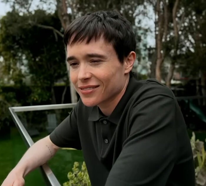 Elliot Page in a dark polo shirt, smiling, sparking controversy regarding biology classes and queer/trans people.
