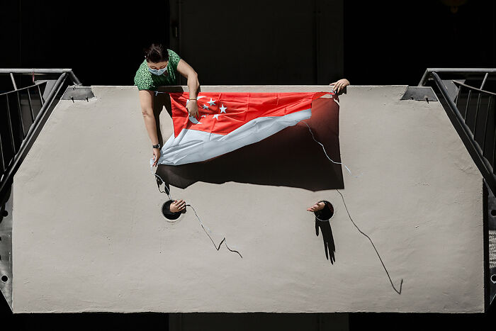 A person holding a Singapore flag while two hands emerge from holes in a wall, for a street photography award.