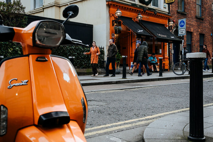 An orange scooter in the foreground, with people walking on a city street in the background, showcasing street photography.