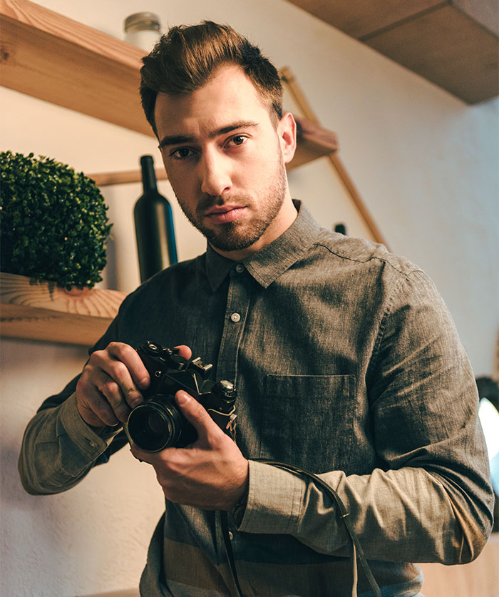A male photographer with a beard, wearing a gray shirt, holds a vintage camera, looking directly at the viewer.
