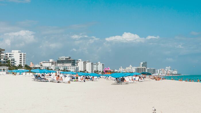 A sunny beach scene with many blue umbrellas and lounge chairs, people relaxing, and a city skyline in the background, illustrating controlling relationships.