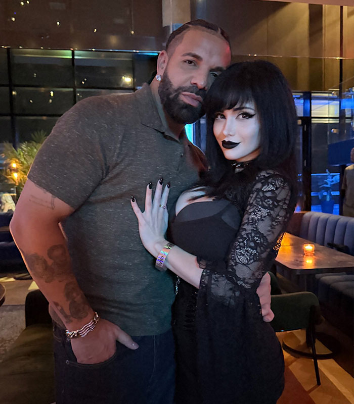 Drake and a Goth Baddie pose together, with the woman wearing a black lace dress and dark makeup.