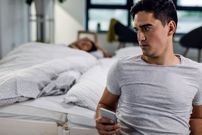 A man looking over his shoulder while holding a phone, with a woman in bed, related to wife material conversations.