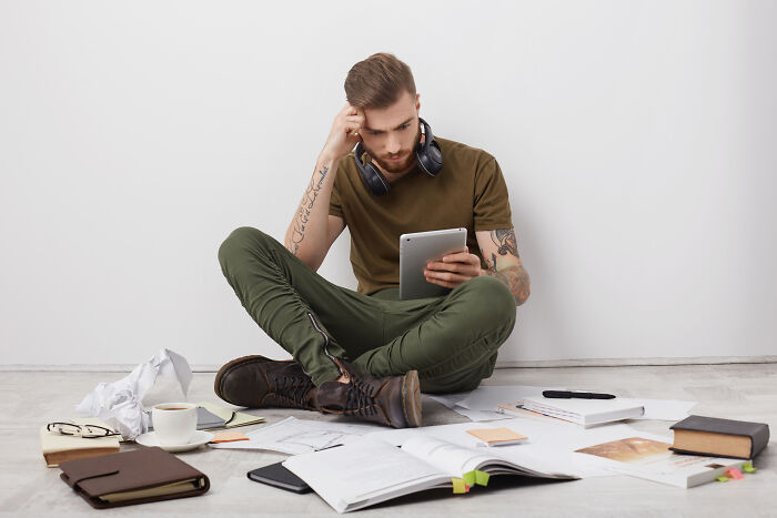 A man with tattoos sitting on the floor surrounded by books and a tablet, highlighting a study routine or habits.