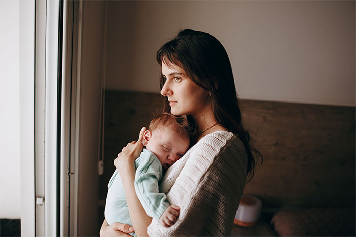 A mother holding her baby with Down Syndrome near a window, looking reflective.