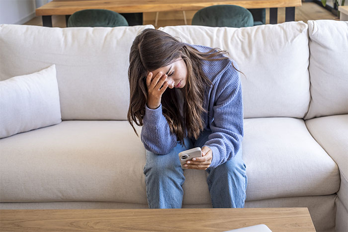 A woman sits on a couch, looking distressed while holding her phone, reflecting on boundaries in a guy's house.