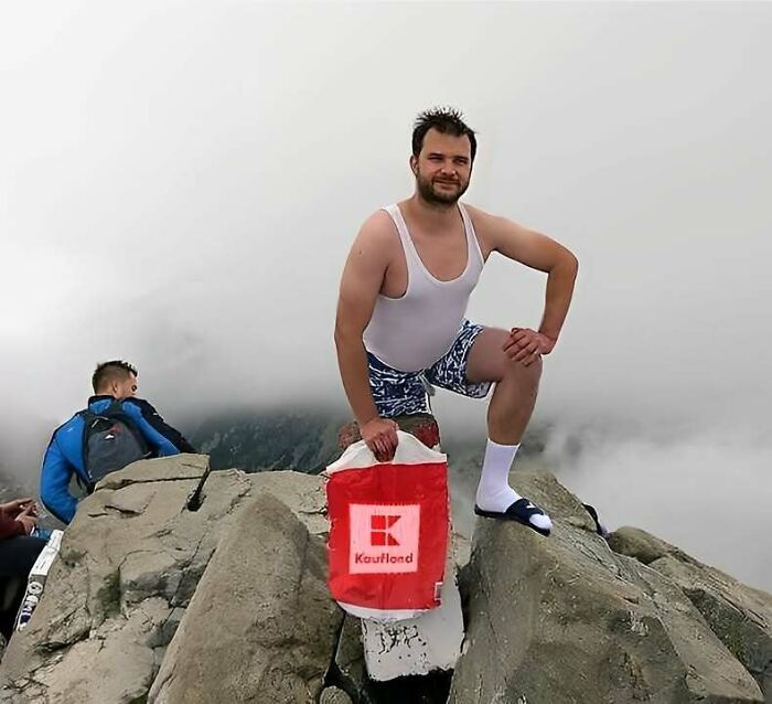 A man in a tank top and shorts posing on a mountain with a shopping bag, showing how Eastern Europe are built different.