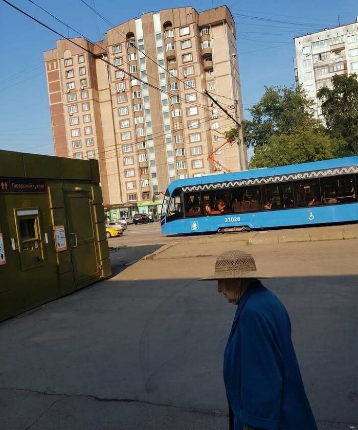 An elderly person in a straw hat walks past a blue tram in a city, highlighting Eastern Europe being built different.
