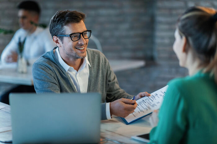 A man in glasses smiling and handing documents to a woman, illustrating employee stories and HR pros.