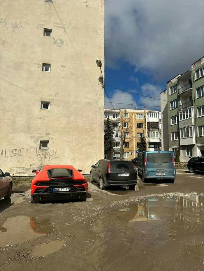 An orange supercar parked in a muddy lot next to old buildings, showing why people from Eastern Europe are built different.