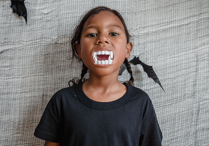 A young, spoiled sister wearing plastic vampire teeth, looking mischievous, like a monster, against a spooky background. A young, spoiled sister wearing plastic vampire teeth, looking mischievous, like a monster, against a spooky background.