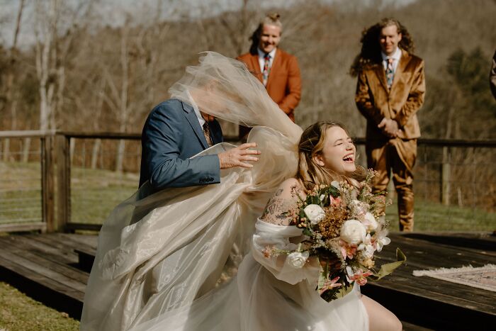 A groom helps a laughing bride whose veil is caught in a funny wedding photo.