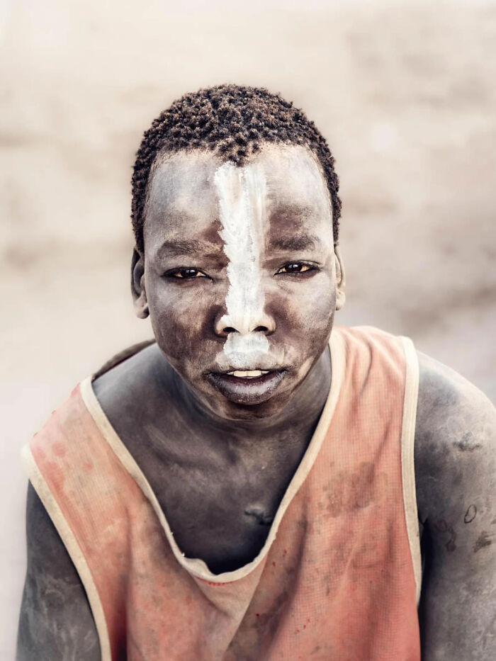 A young boy with a dirt-covered face crouches on the ground, creating shapes from mud. This image won a 2026 European Photography Award.