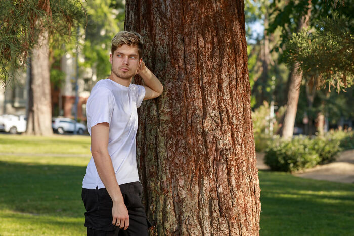 A young man reveals a secret while leaning against a tree in a park, looking thoughtfully to the side.