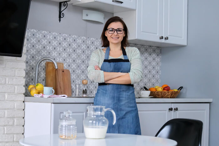 A smiling woman in an apron and glasses standing in a modern kitchen, related to dumb reasons couples divorce.