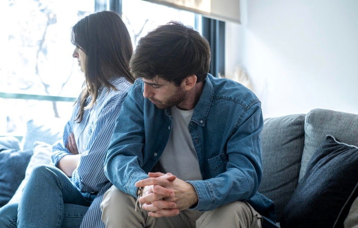 A man questioning marriage, looking down with folded hands while his wife sits with her back to him, angry.