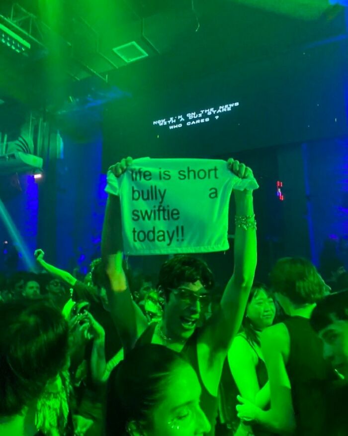 Iconic shirts: Person at a concert holding up a white shirt that reads, 'life is short bully a swiftie today!!'