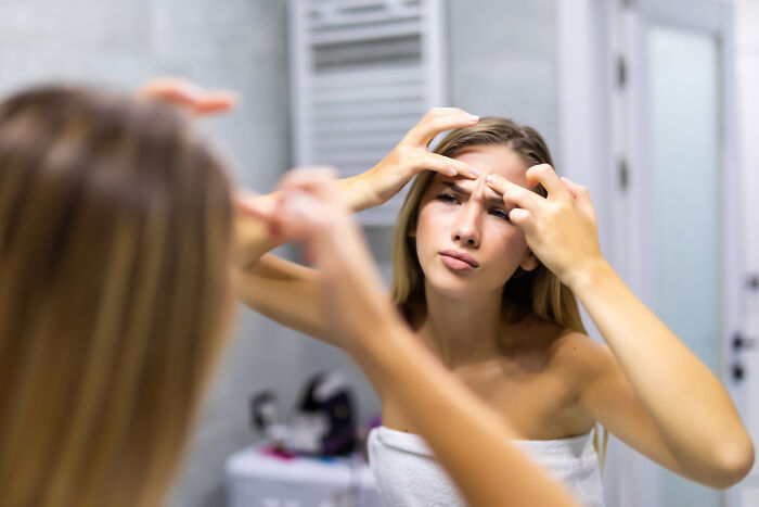 A young woman in a towel looks into a mirror, picking at her face. This is something doctors refuse to do.