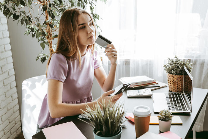 A thoughtful woman holding a credit card and phone, sitting at a desk with a laptop, highlighting skills that change lives.