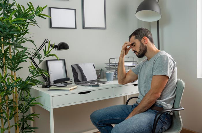 A man sits at a desk, head in hand, looking stressed, illustrating the impact of dumb work rules.